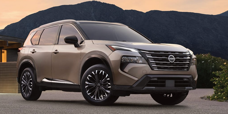 A gold Nissan Rogue SUV with a black roof and black wheels at sunset with mountains in the background.