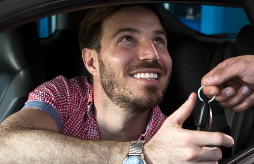 A man in a red checkered shirt smiling as he receives car keys from another person's hand.
